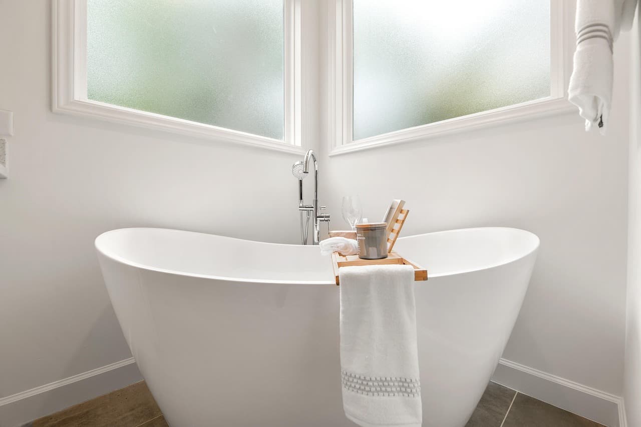 A white freestanding bath under a window in a pale painted bathroom
