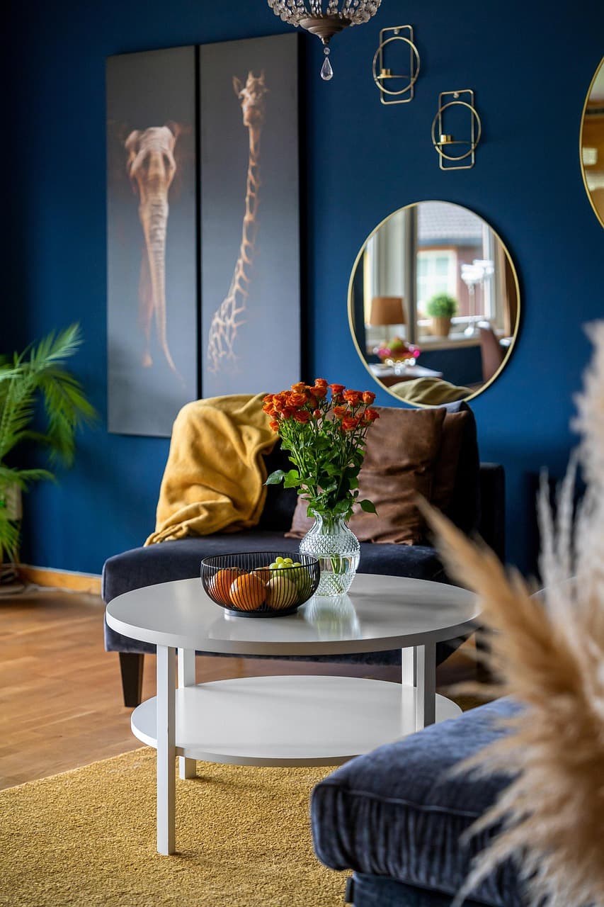 A living room painted deep blue, with a round mirror, a yellow throw and flowers